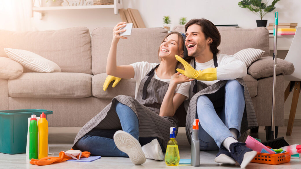 Vysmiaty mladý pár oddychuje s chytrým telefónom po jarnom upratovaní bytu.