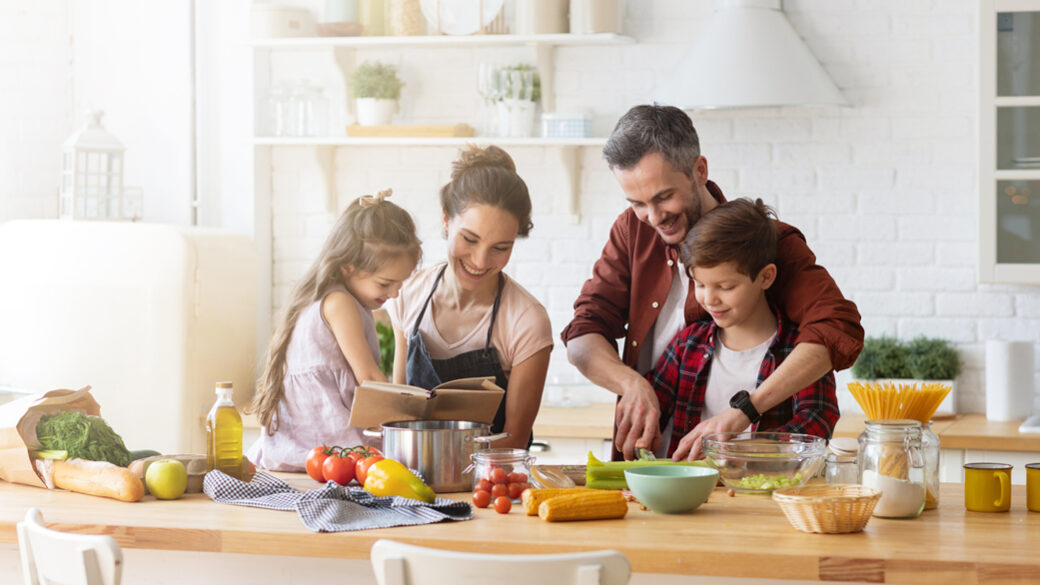 Šťastná rodina společně vaří v kuchyni. Matka s dcerou čtou recept tatínkovi a synovi.