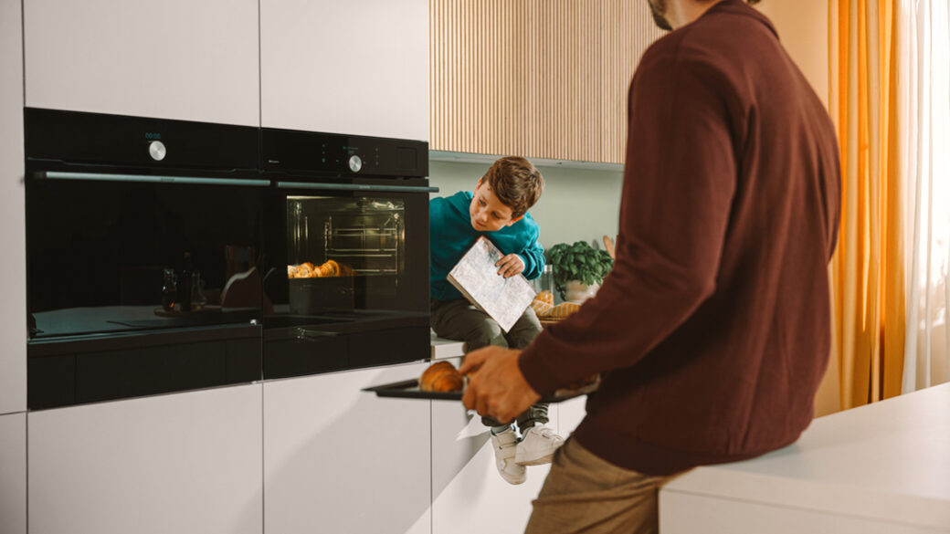 A man taking croissants out of a modern built-in Gorenje G800 oven while a young boy watches with a smile, sitting on the kitchen counter holding a book. Warm, natural light fills the stylish kitchen.