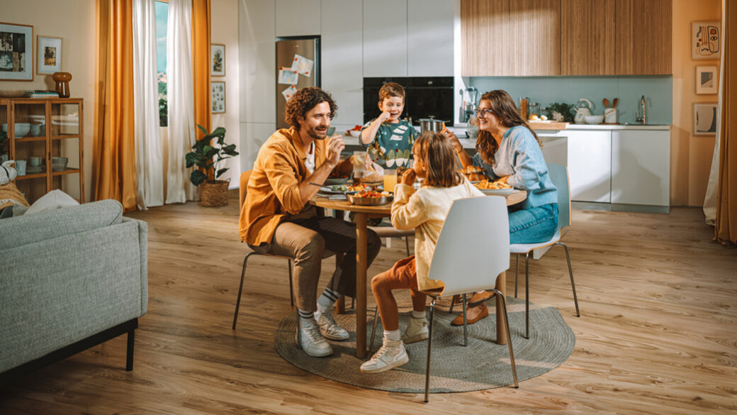 A young family of four enjoying a cheerful meal together at a round kitchen table in a bright, modern home. The parents and two children are smiling and chatting, surrounded by warm wooden tones and soft natural light.