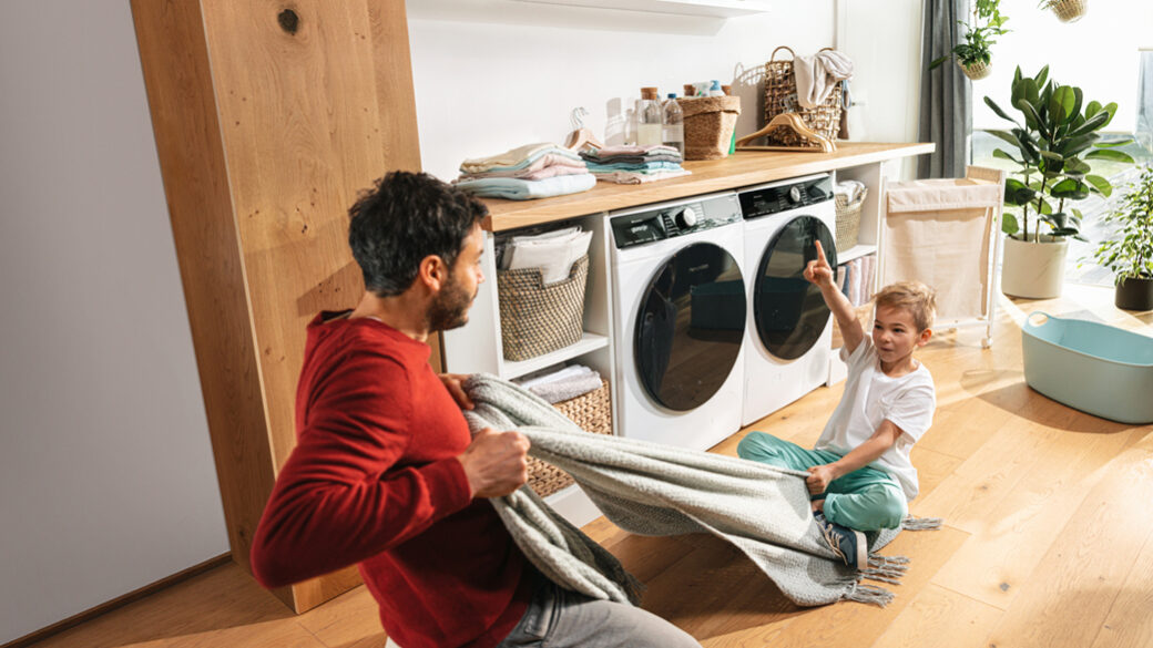 A father and young son playing together in a bright, modern laundry room. The child is sitting on a blanket being gently pulled, laughing and pointing upward, while the father smiles.