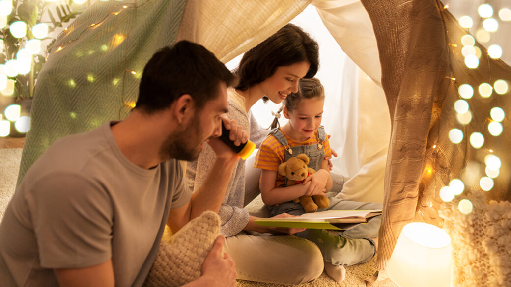 A family sitting together in a cozy blanket fort lit with warm string lights, reading a book and smiling.