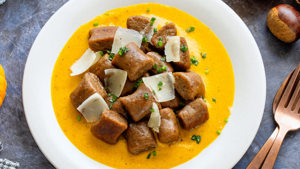 A plate of chestnut gnocchi served in a creamy orange sauce, topped with shaved cheese and chopped herbs, styled with autumn elements like chestnuts and small pumpkins.