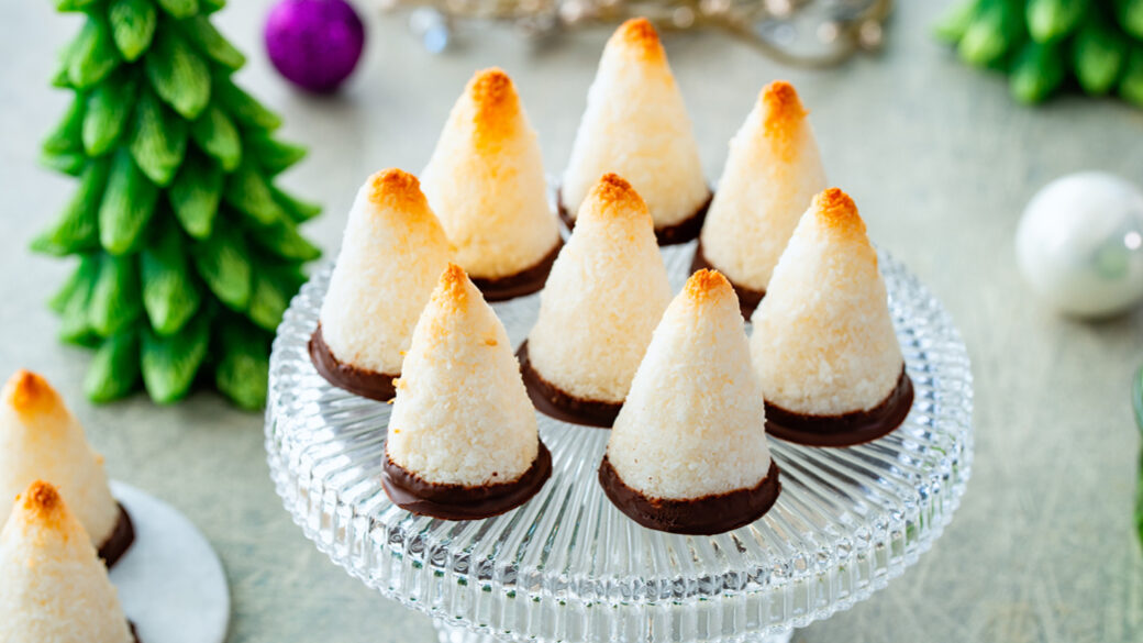 Chocolate-dipped festive coconut macaroons shaped like small Christmas trees on a glass serving stand.