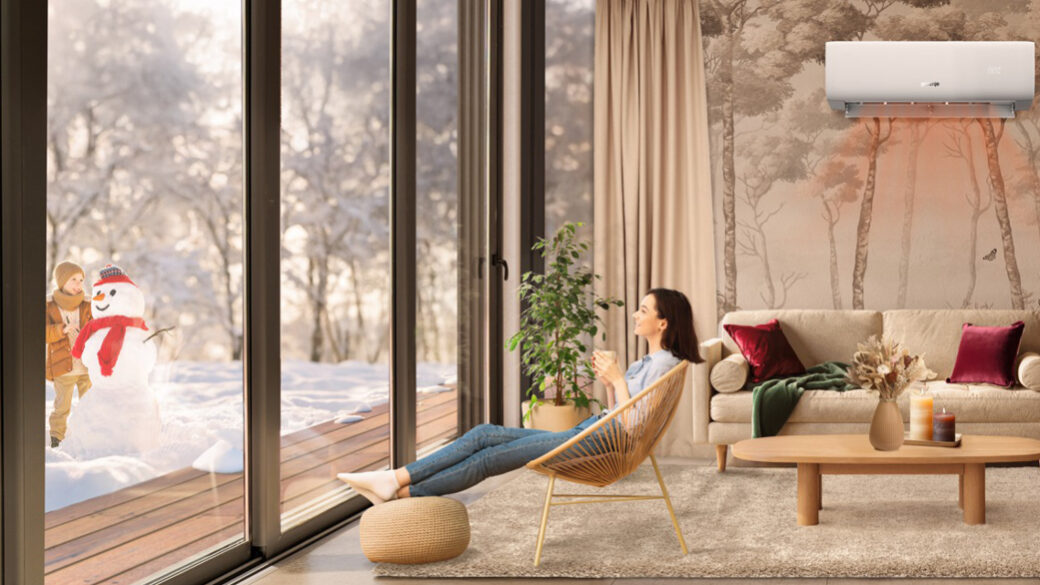 Woman relaxing with a warm drink in a cosy living room heated by a Gorenje air conditioner, while a child builds a snowman outside in a snowy winter landscape.