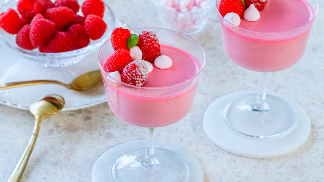 Two glasses of raspberry and rose posset topped with fresh raspberries and mini meringues, served on a light stone surface with gold spoons and a bowl of berries.