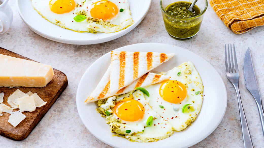 A breakfast plate with sunny-side-up eggs cooked in basil pesto cream, garnished with fresh herbs and chilli flakes, served with toasted tramezzini bread.