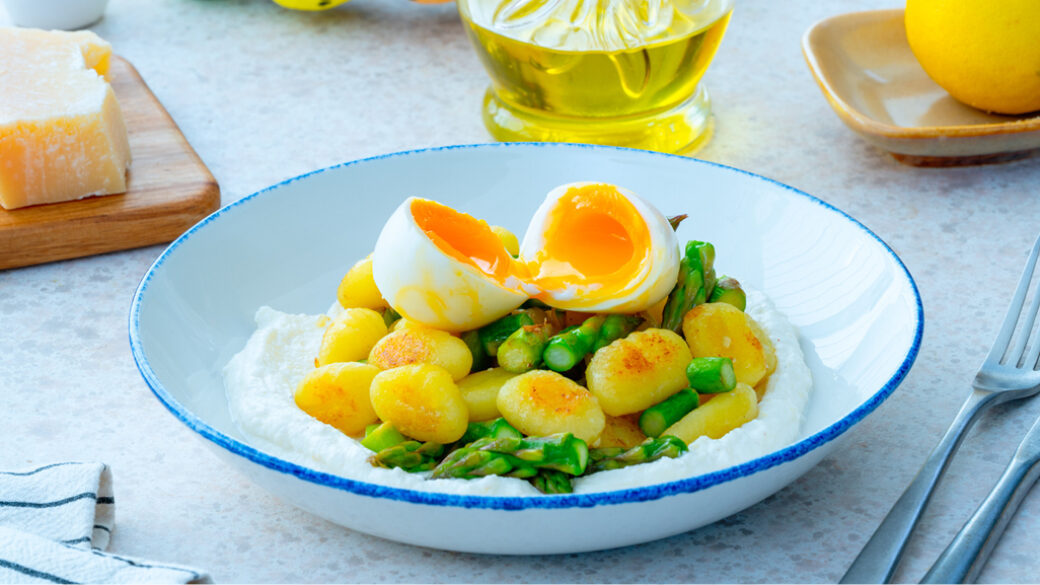 Crispy golden gnocchi and fresh asparagus served over a bed of whipped ricotta, topped with a soft-boiled egg sliced in half to reveal a jammy orange yolk, in a white bowl with a blue rim.