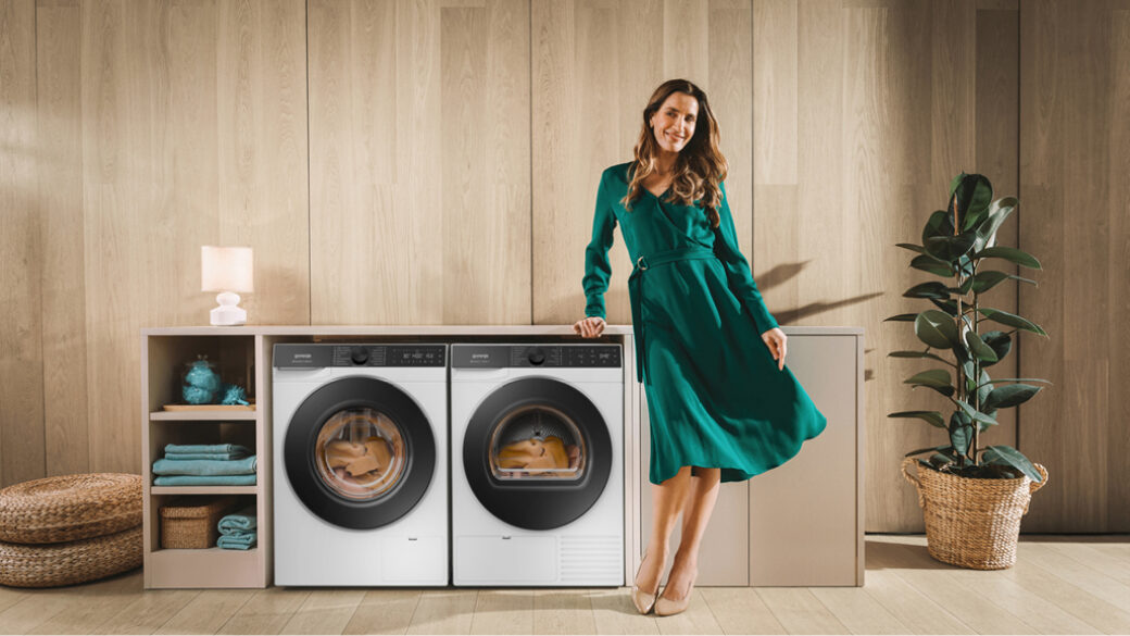 A woman in a green dress standing beside a Gorenje SimpliCare washing machine and tumble dryer in a modern, minimalist laundry space with wooden walls and soft lighting.